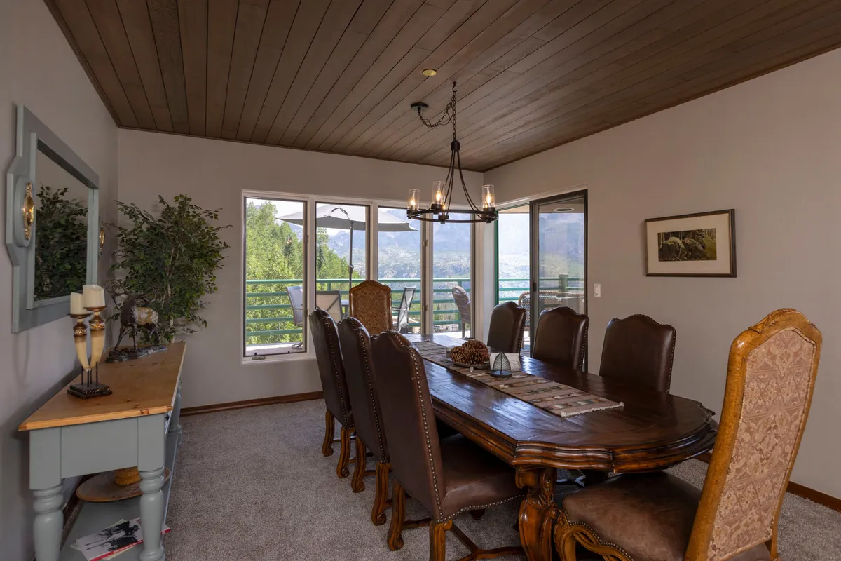A large wooden dining table under a stylish chandelier, flanked by windows that offer scenic views. A decorative mirror and a touch of greenery enhance the cozy, welcoming atmosphere, making it ideal for memorable meals and gatherings.