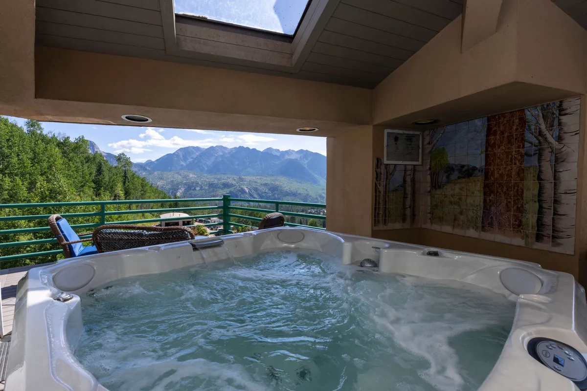 A serene lounge area with a private hot tub poised under a skylight, offering spectacular views of the surrounding mountains. There is comfortable seating nearby, and the scene is bathed in natural light, emphasizing the connection to the outdoors and the tranquility of the setting.