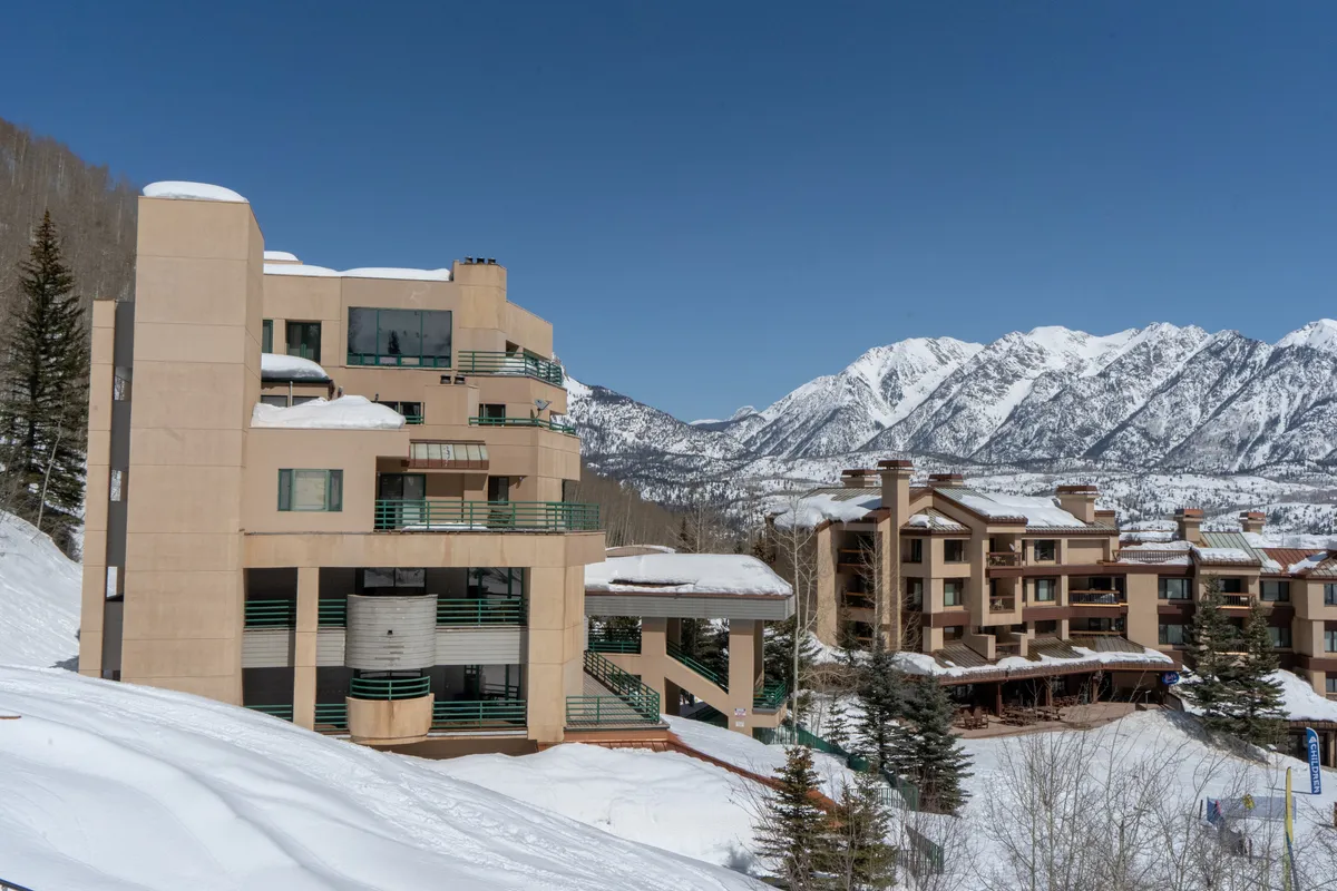 A sweeping exterior view featuring a mountain range with snow-covered buildings nestled below and pine trees dotting the landscape. The scene evokes a sense of adventure and serenity, suggesting proximity to ski slopes and mountain exploration.