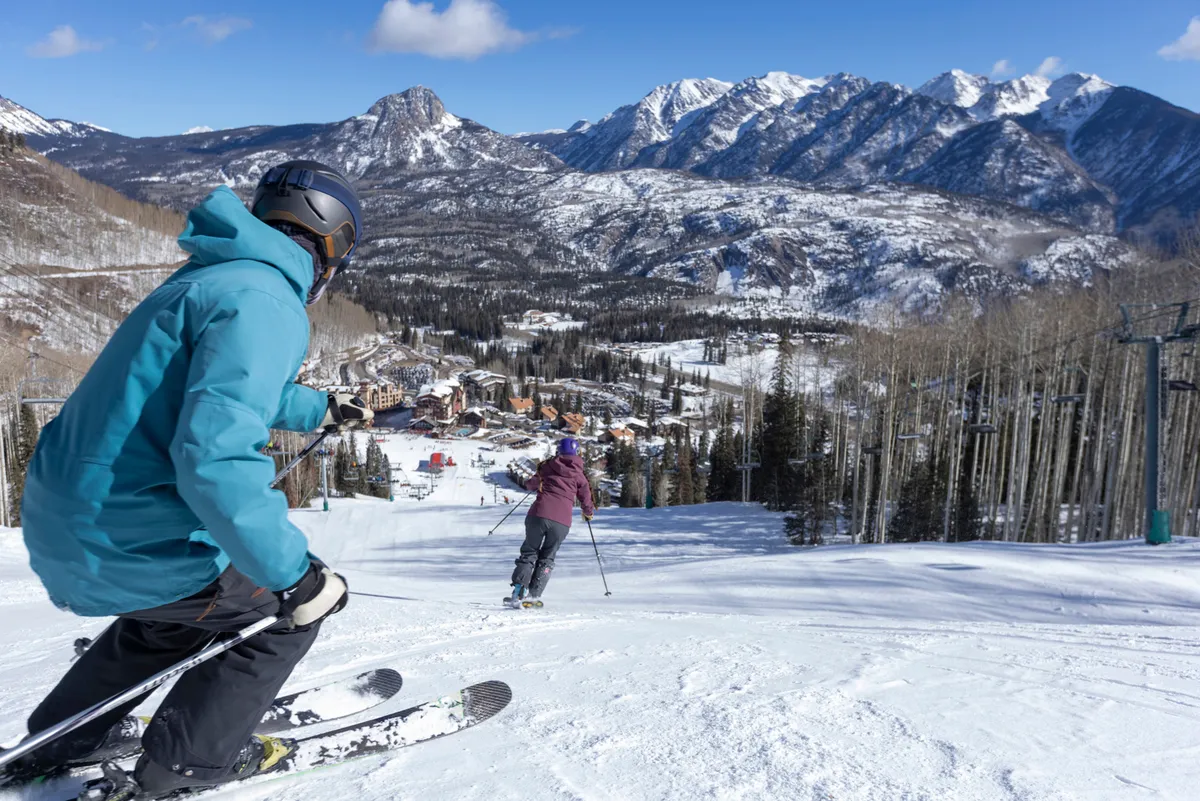 Skiers descend a snowy slope with towering mountains in the background and a ski resort nestled in the valley below. The action and energy of the scene underline winter sports and shared outdoor excitement.