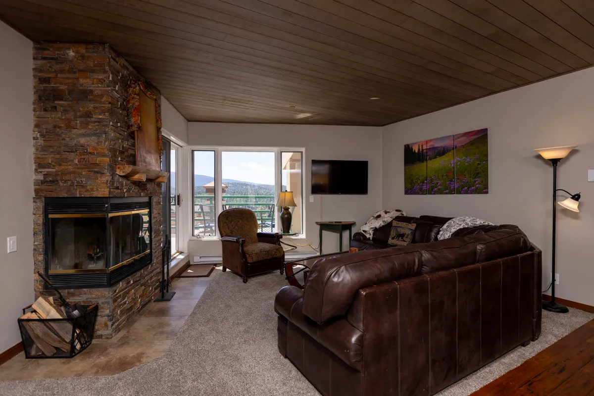 A comfortable open-concept living room with a stone fireplace, generous seating, large windows framing outdoor views, and a television. The space feels warm, relaxed, and visually connected to adjoining areas.