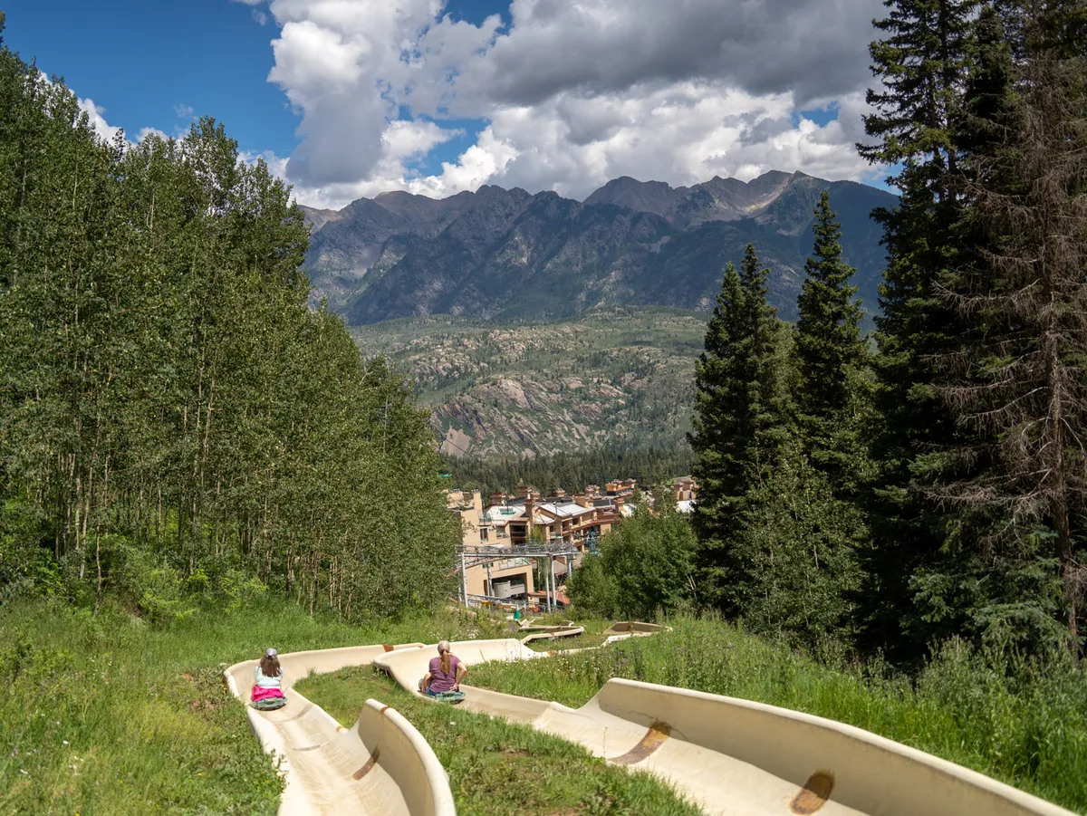 A lively scene showing people enjoying a summer mountain slide, with lush greenery, expansive mountain views, and the village below. The image captures excitement and adventure, highlighting the vibrant atmosphere surrounding the condo.