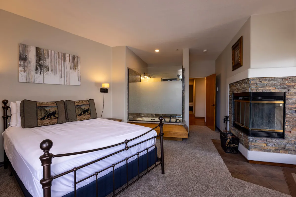 This image showcases a bedroom with a queen-sized bed adorned with decorative pillows, a cozy fireplace, and a glass partition leading to a bathroom. The soft lighting and warm tones create a tranquil and comfortable atmosphere perfect for relaxation.