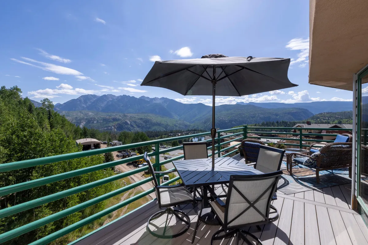 A spacious outdoor dining area featuring a large table with an umbrella, surrounded by lounge chairs and overlooking majestic mountain views. The inviting setting is perfect for group meals, celebrations, or simply relaxing while taking in the scenery.
