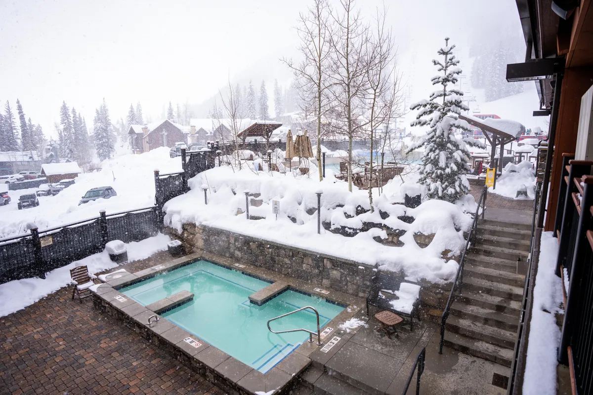 An inviting hot tub set amidst a snowy landscape, framed by evergreen trees and stone steps. The image portrays guests unwinding in the warm water, creating a cozy, social scene with a backdrop of winter scenery.