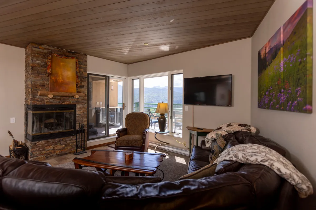 A welcoming living room featuring a central fireplace, expansive comfortable seating, and large windows framing dramatic mountain vistas. The warm glow from the fire creates a cozy environment perfect for relaxing or socializing, while the view through the windows captures the alpine landscape and ski slopes.
