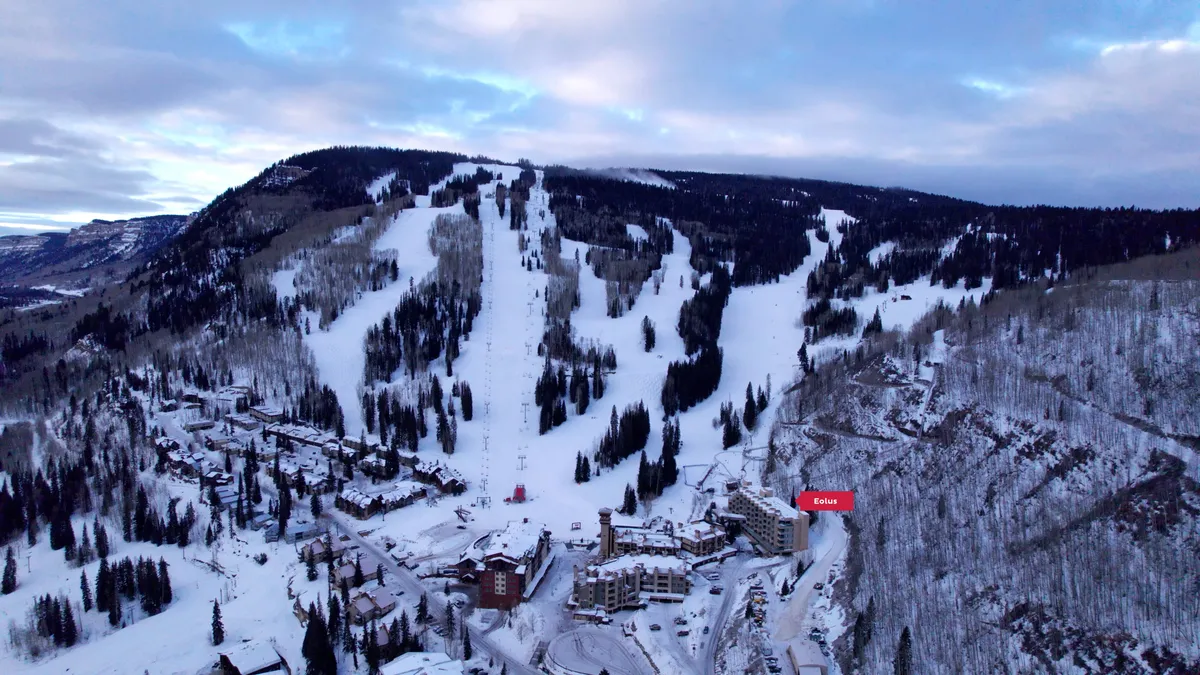 A panoramic exterior view of Purgatory Resort with the ski slopes in the foreground, surrounded by a snowy pine forest and resort buildings nestled in the landscape. The snow-covered terrain is bustling with skiers and snowboarders, capturing the winter adventure atmosphere.