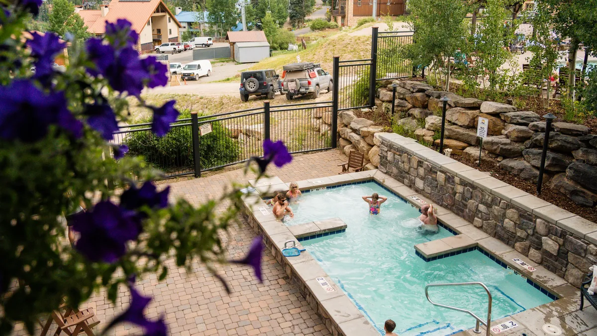 This image features the outdoor swimming pool surrounded by lush landscaping and people relaxing, suggesting a vibrant yet relaxing resort atmosphere. The flowers in the foreground, clear blue water, and people sunbathing or socializing capture the leisurely, energetic spirit of resort living.