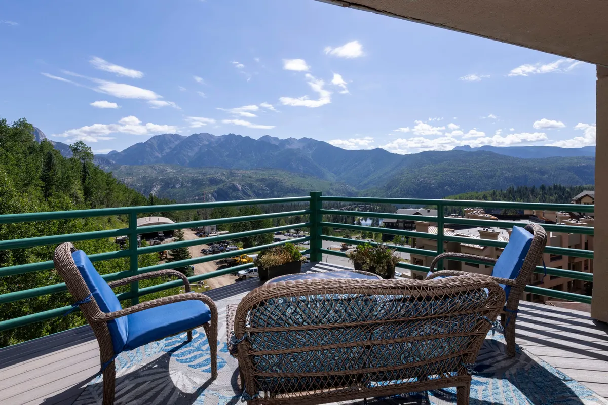 The hero image showcases a private outdoor deck with comfortable seating surrounded by potted plants, set against a breathtaking backdrop of the Needles Mountains. The scene captures the serene atmosphere perfect for relaxing or socializing while taking in expansive mountain vistas.